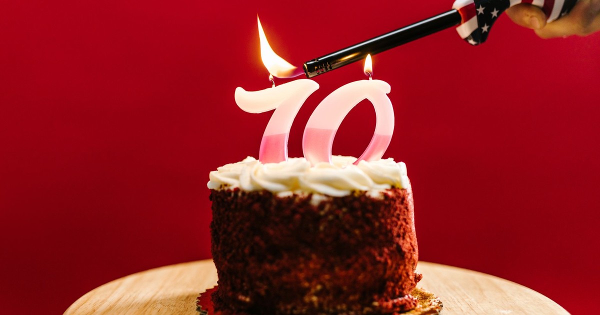 A close-up of a birthday cake with '70' candles being lit against a red background.