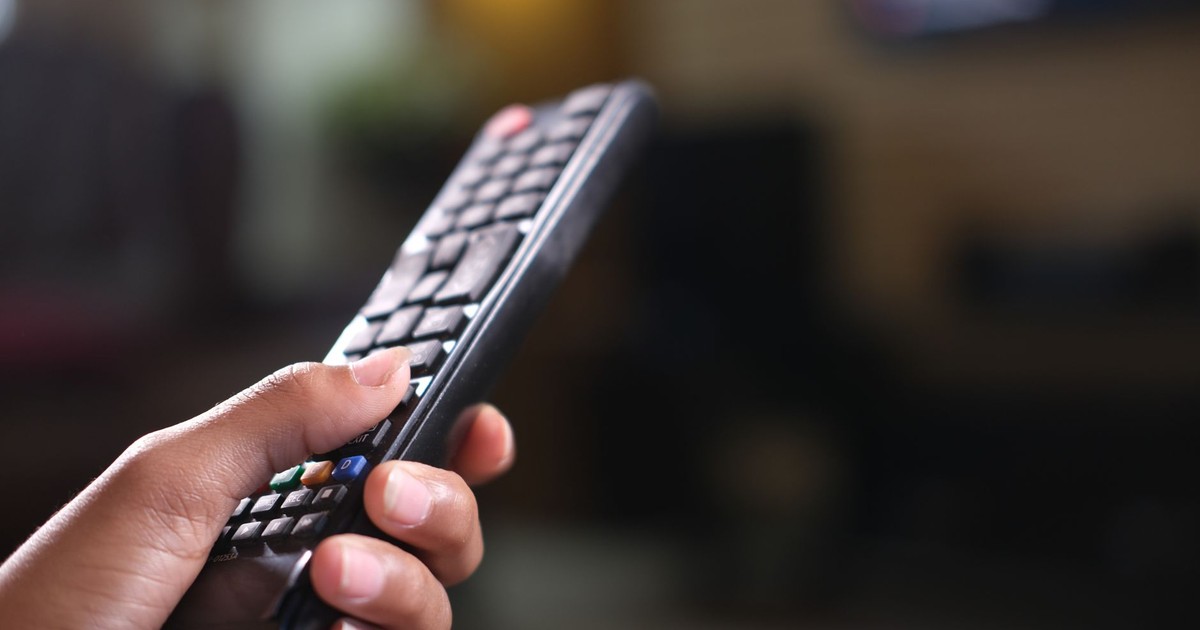 Hand holding a TV remote control, focusing on buttons and grip indoors.