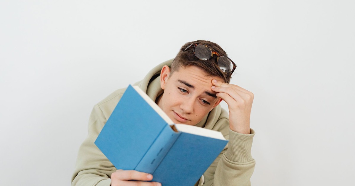 Teenager reads a book while holding glasses on his head, against a plain white background.