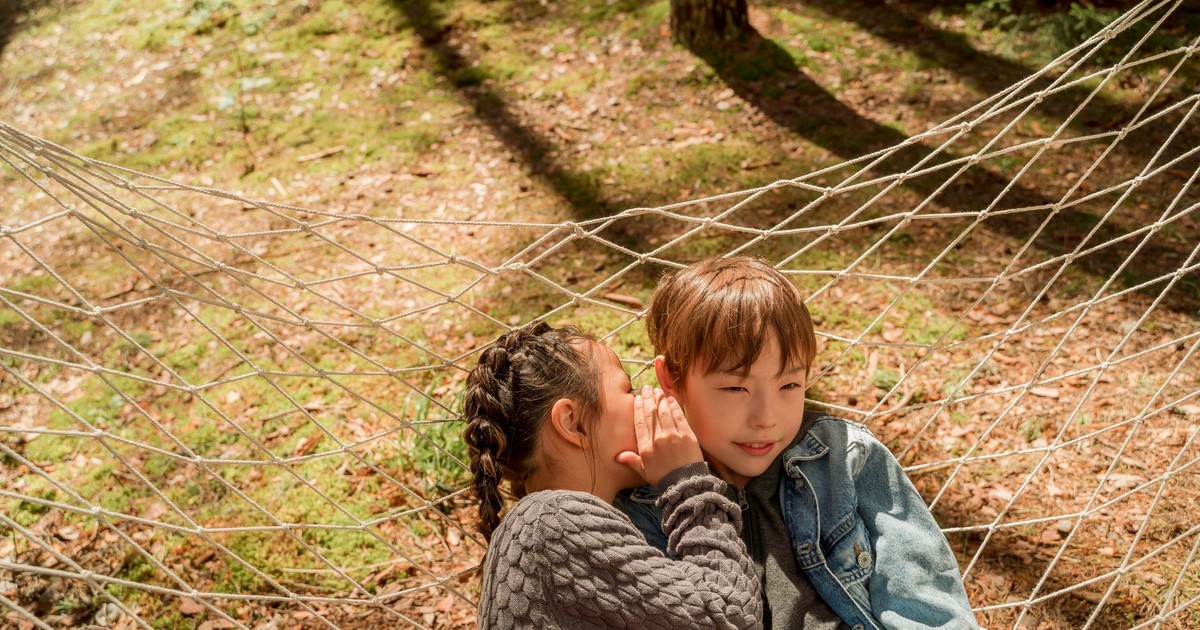 Two children sharing a secret while sitting in a hammock in a sunny outdoor setting.