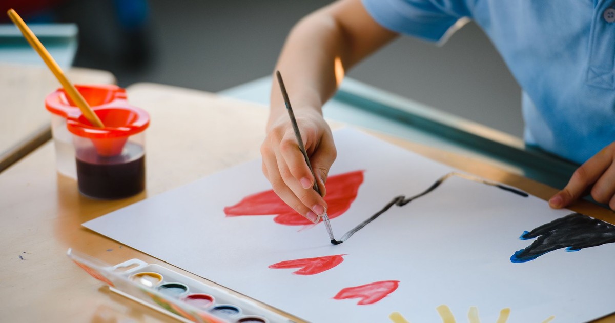 Young child using watercolors and brush to paint on paper indoors at a school setting.