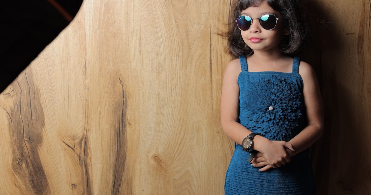 Cute and fashionable young girl wearing sunglasses and a blue dress against a wooden backdrop.