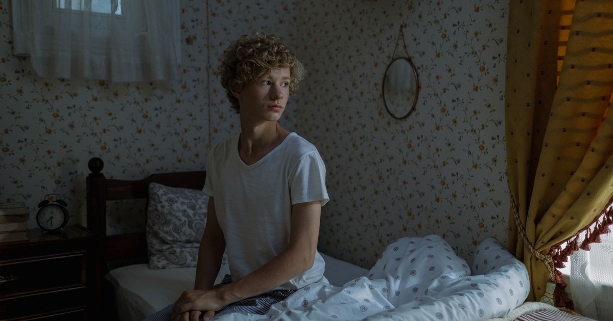 Teenage boy with curly hair sitting on a bed in a cozy, dimly lit bedroom.