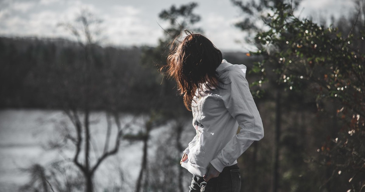 A woman in a white hoodie stands near a lake surrounded by nature during the day.
