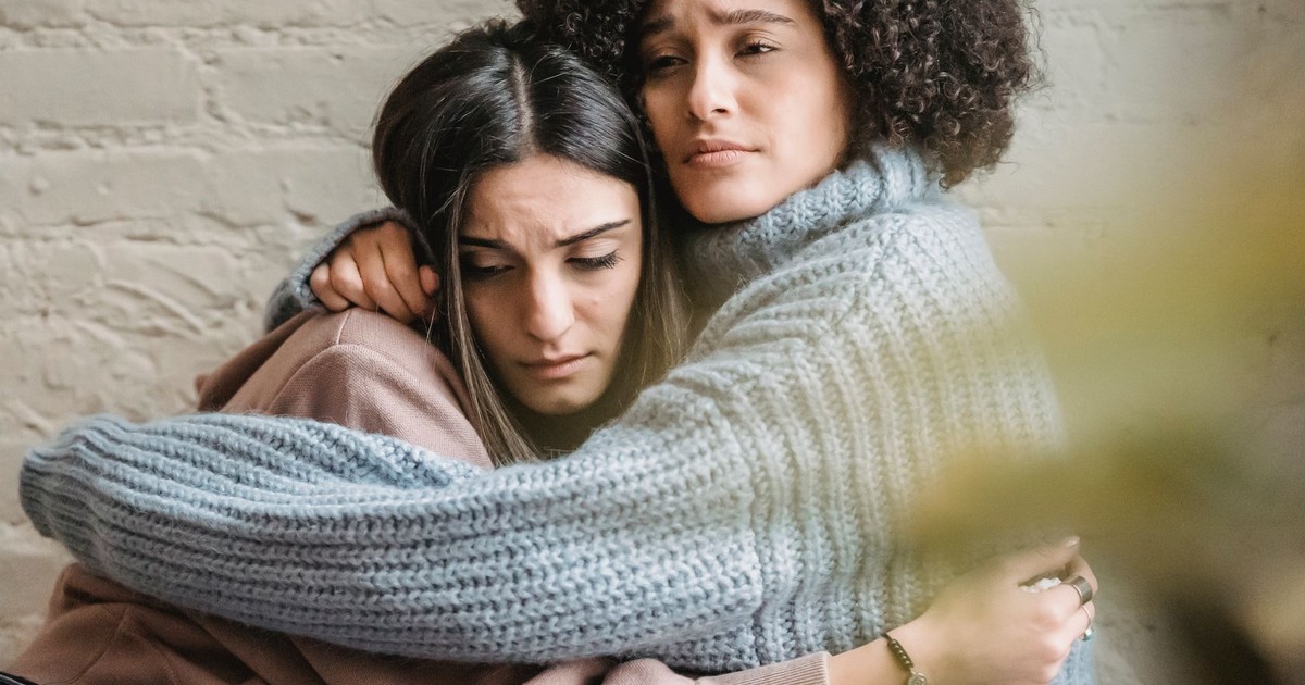Sad multiracial female friends in casual clothes embracing while sitting near wall in light room with green plant at home