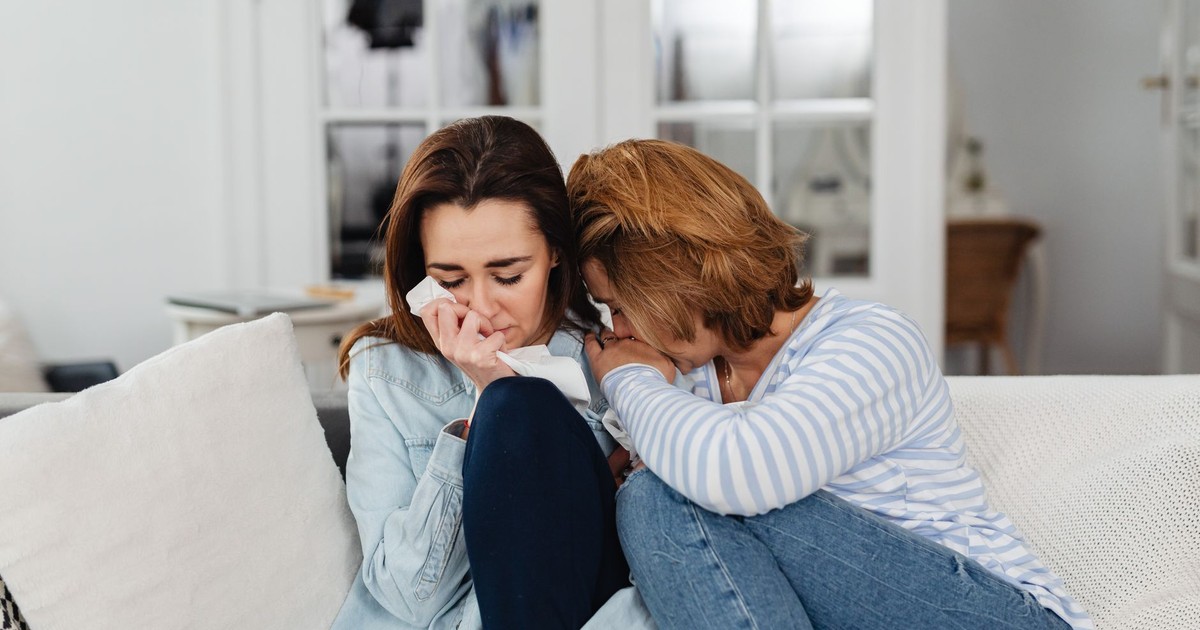 Two women sitting on a couch, comforting each other at home, displaying emotions of sadness and support.