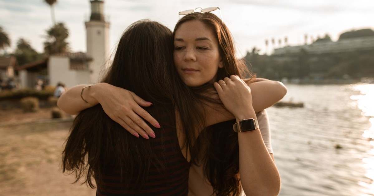 Two friends share a heartfelt hug beside a serene lakeside with a lighthouse in the background.