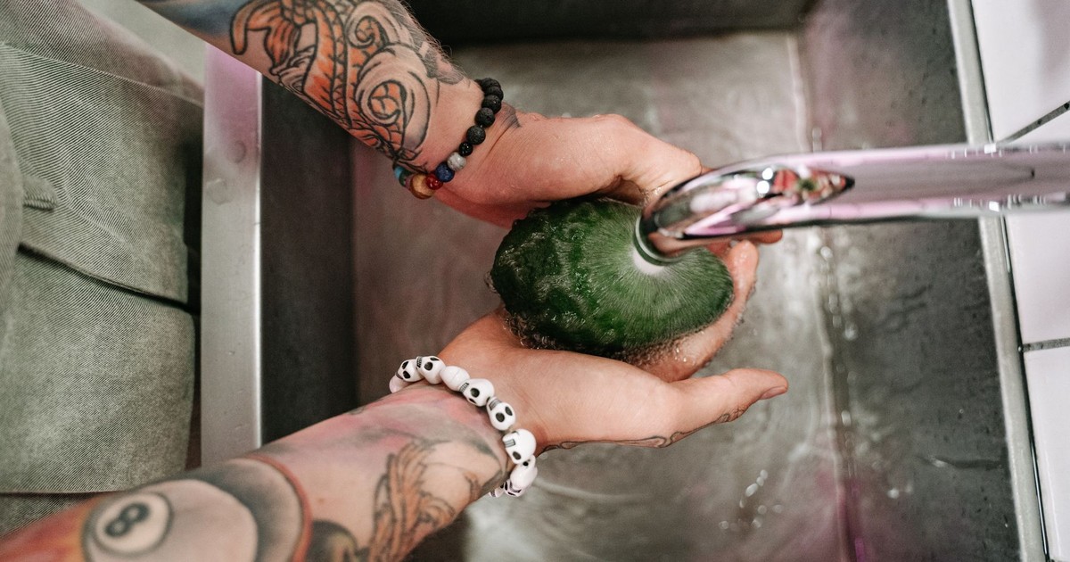 Top view of tattooed hands washing an avocado under running water in a kitchen sink.