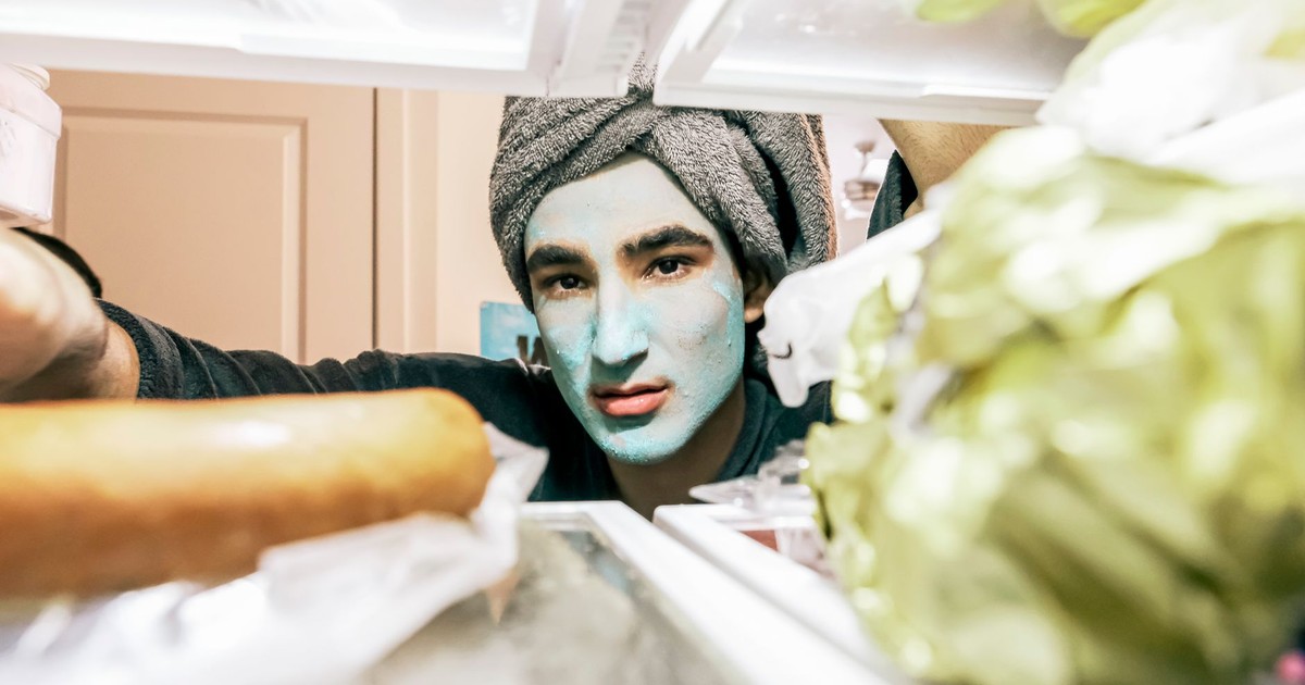 A man with a face mask peers into a refrigerator, wrapped in a towel.