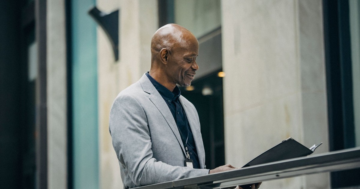 SIde view of middle aged smiling black executive in formal suit standing near fence and reading papers in card board near modern office