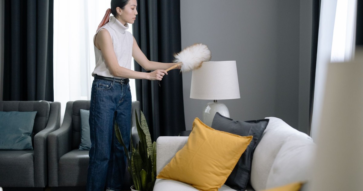Asian woman dusting a lampshade in a modern living room with yellow pillows.