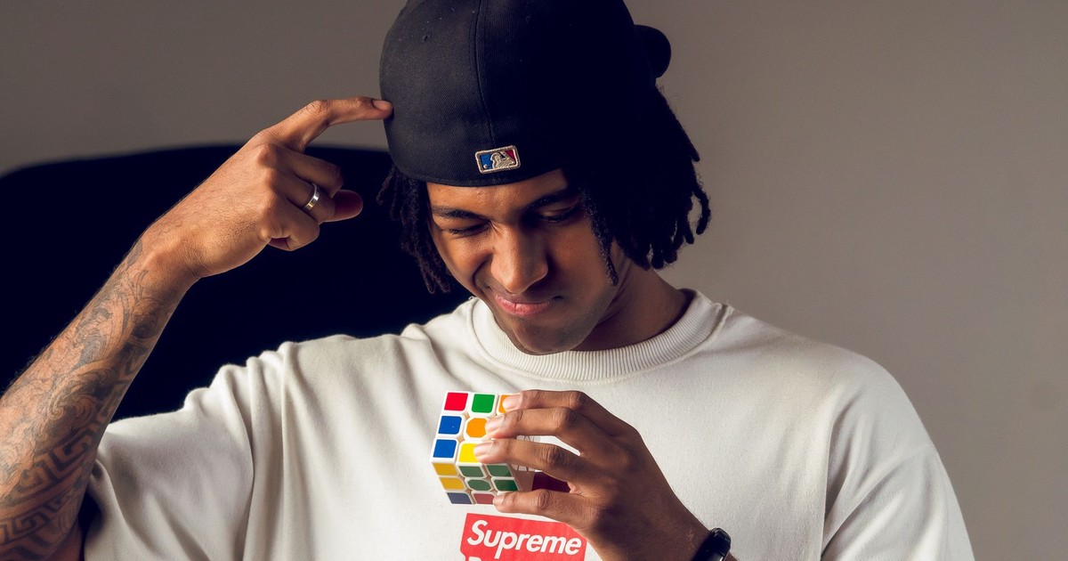 A thoughtful young man wearing a cap focuses on solving a colorful puzzle cube indoors.
