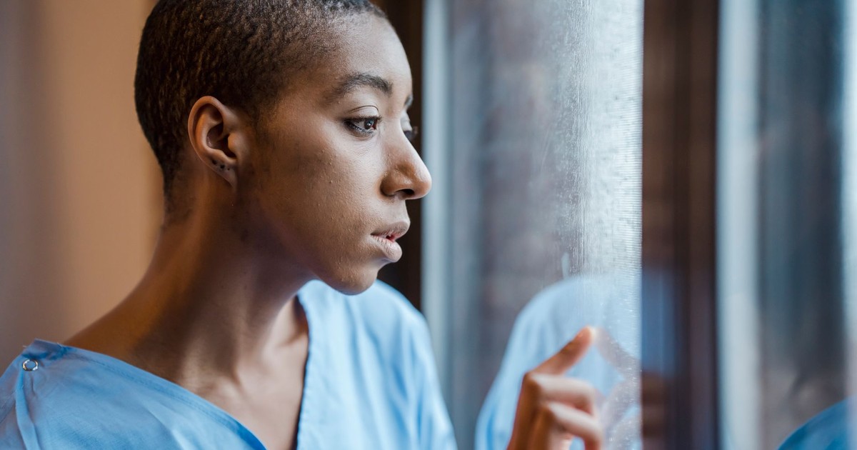 Upset African American female patient in medical wear touching window while looking away with concern