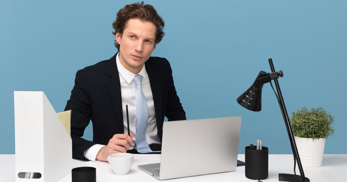 A focused businessman in a suit works at his tidy desk with a laptop, plant, and lamp.