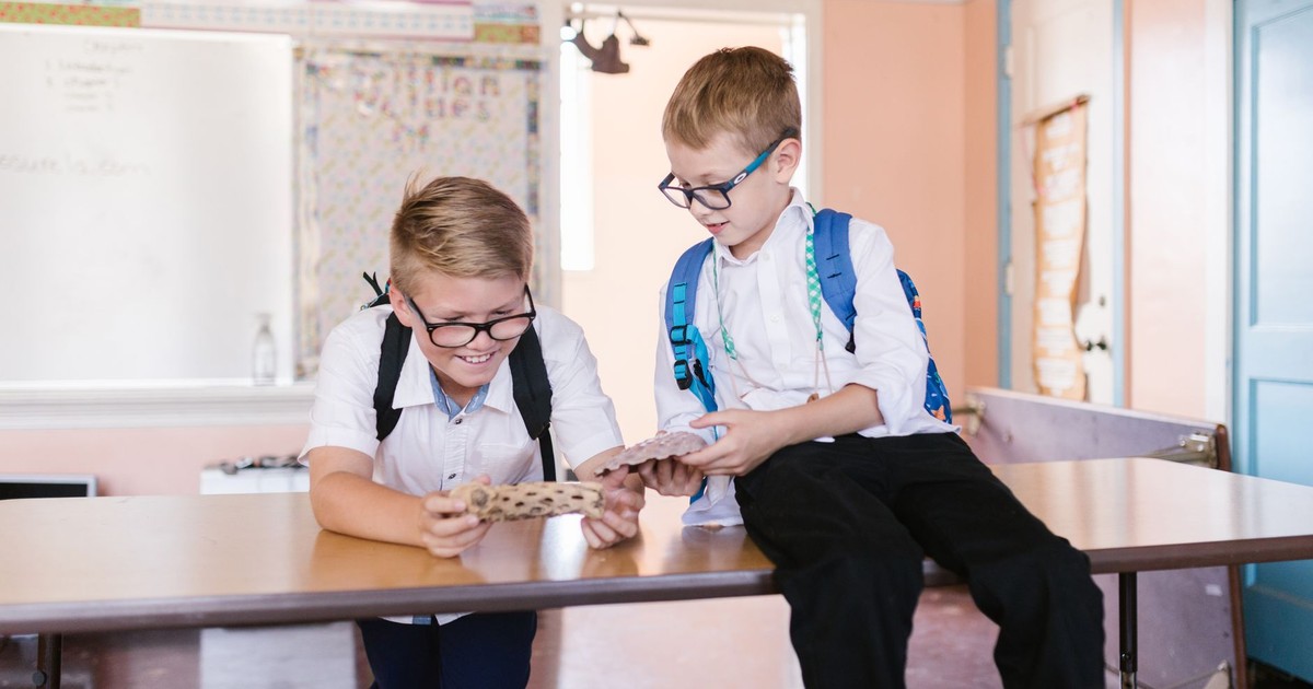 Two boys in school uniforms laughing and looking at a mobile phone in a classroom.
