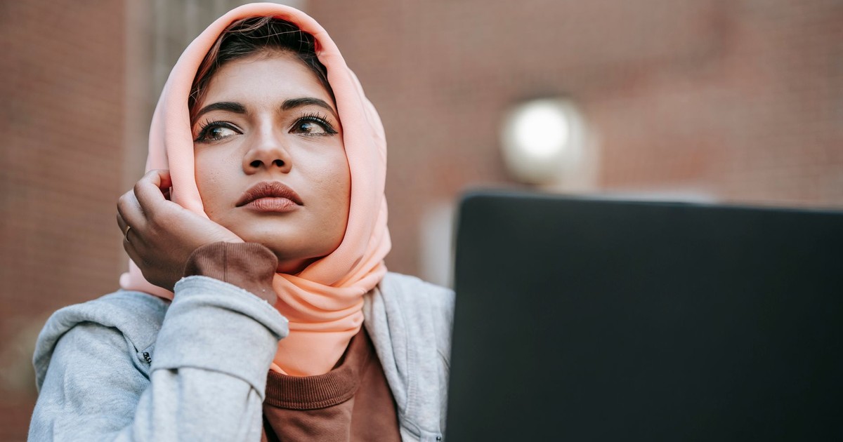 Dreamy young Muslim female student wearing hijab sitting with portable laptop and leaning on hand while looking away in daylight