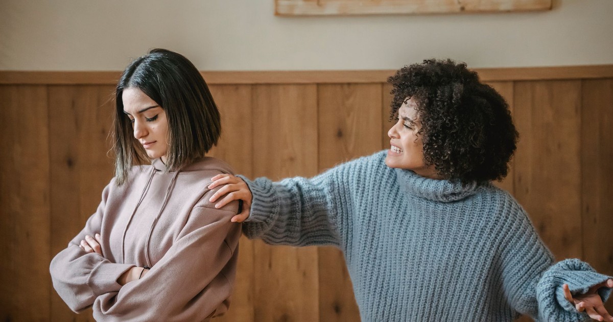 Unhappy young multiethnic female friends in casual outfit arguing with hand on shoulder while standing in bright house near wall