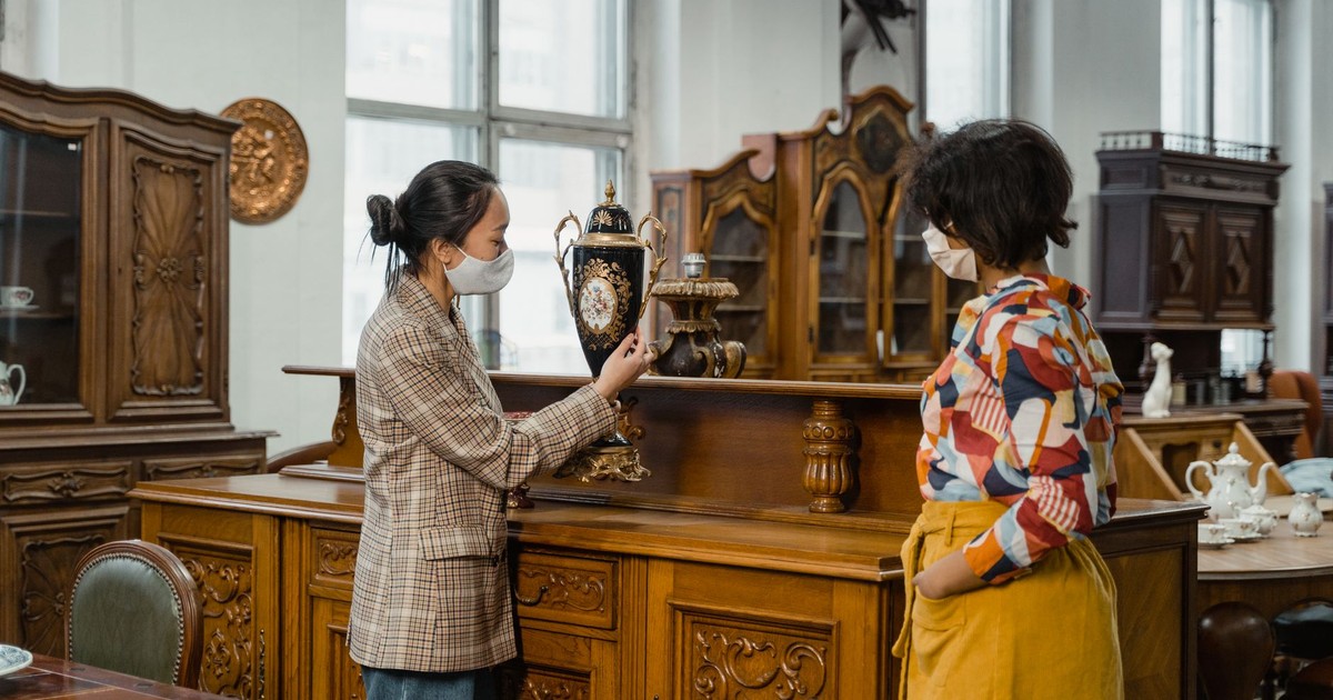 Two women wearing masks browse an antique store, examining a decorative vase.