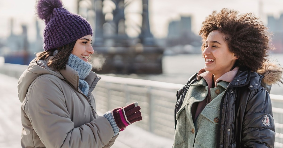 Joyful young multiracial female friends in warm clothes chatting happily and looking at each other with smiles while standing on city embankment on sunny winter day