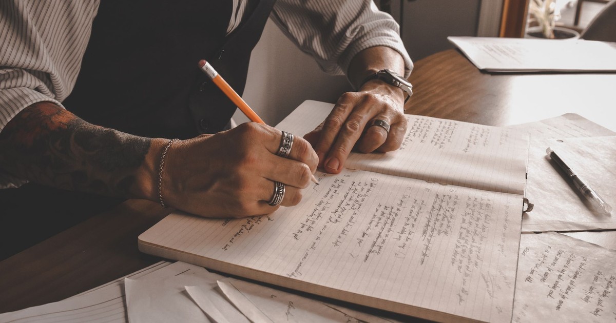 Close-up of a tattooed hand writing in a notebook with pencil and papers around.