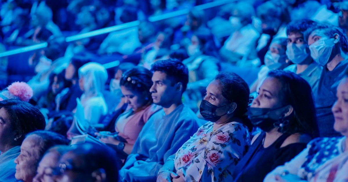 A diverse audience in masks attentively watching a concert under blue lights.