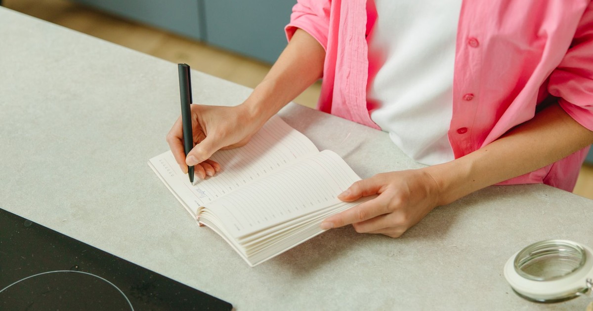 A woman in a pink shirt writing in a notebook on a kitchen counter with a pen.