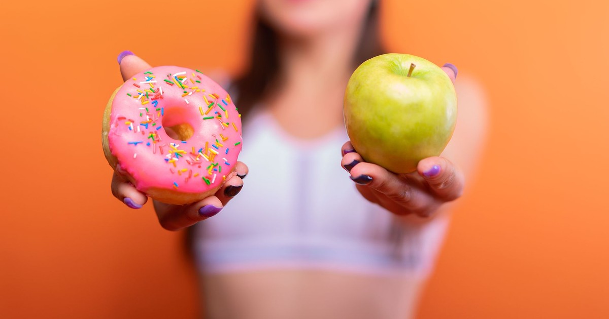 A woman holds a green apple and a pink donut with sprinkles against an orange background, emphasizing choice.