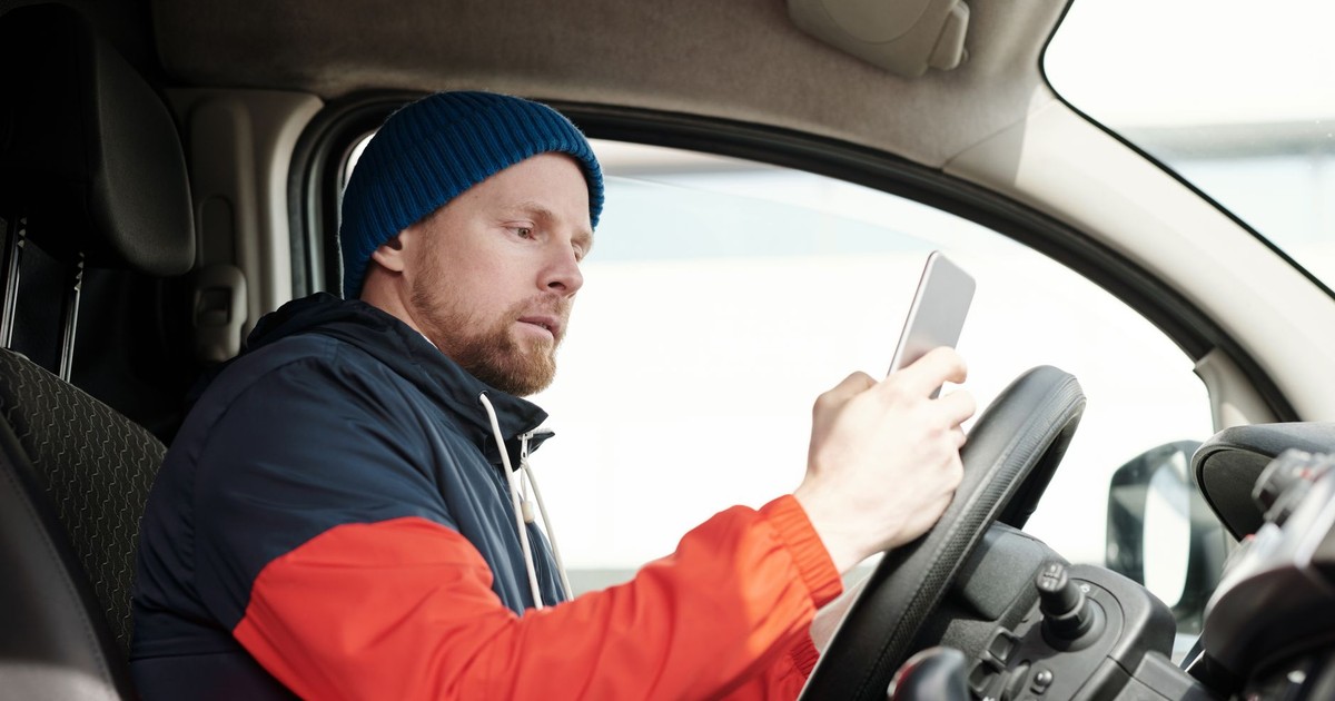 Man in a parked car using a smartphone, wearing a blue beanie and red jacket. Daytime driving scene.