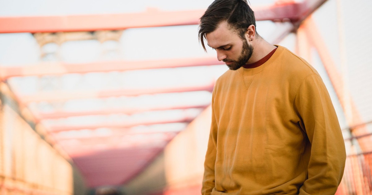 Young thoughtful bearded male in casual apparel with modern haircut looking down while standing with hands on hips in town