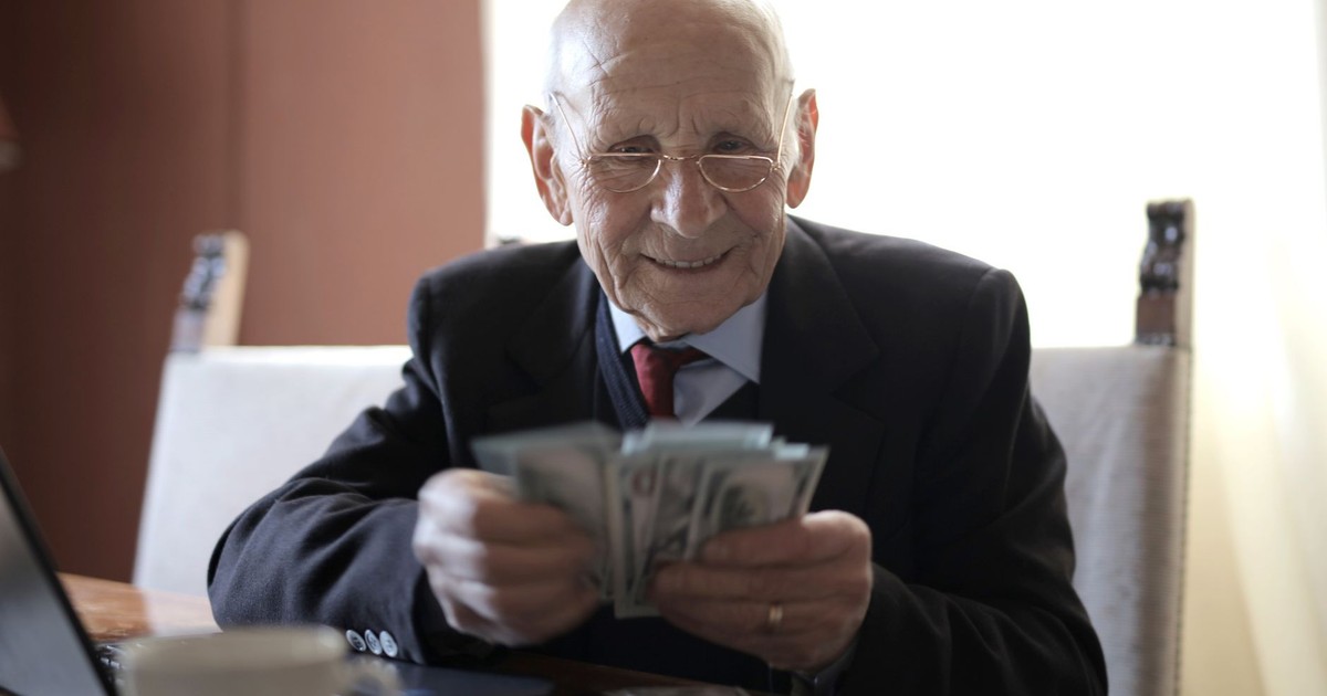 Positive senior businessman in formal suit and eyeglasses counting money bills while sitting at wooden table with cup of beverage and near opened laptop