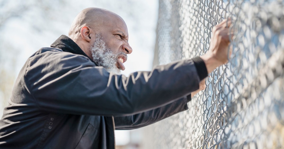 A frustrated man passionately grips a chain-link fence, expressing stress and determination.