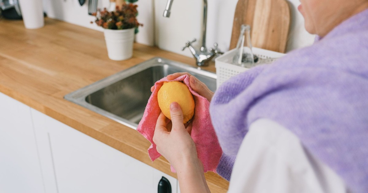 Crop unrecognizable person in casual clothes standing and wiping ripe fresh lemon with cloth in light kitchen in daytime