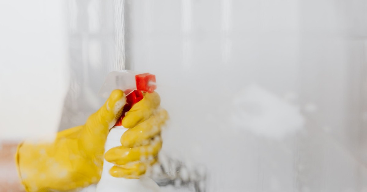 Crop unrecognizable person in yellow latex protective glove spraying detergent from bottle with plastic dispenser at glass of shower cabin