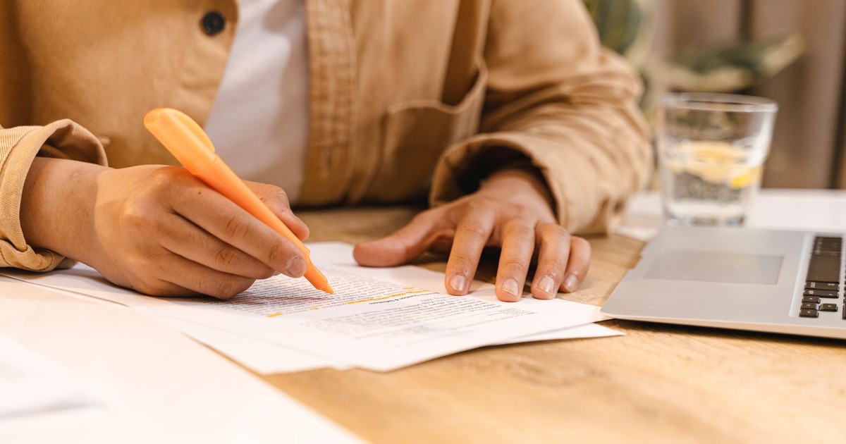 Person highlighting notes at a desk with a laptop and glass of water nearby.