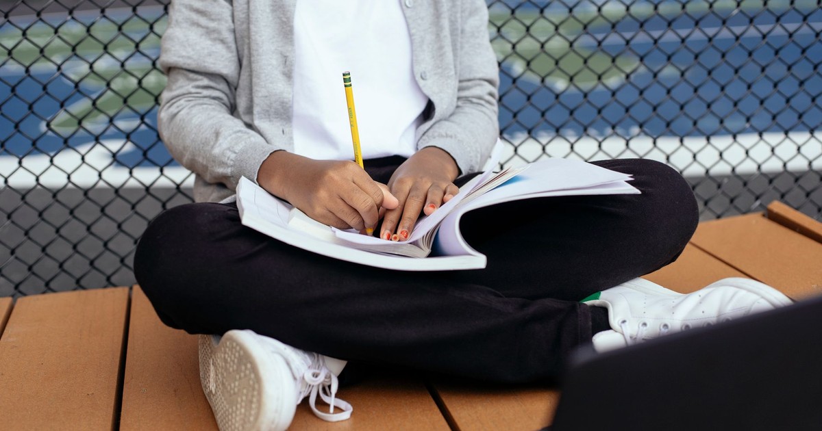 Crop unrecognizable schoolkid sitting with crossed legs and writing notes in copybook while studying with laptop