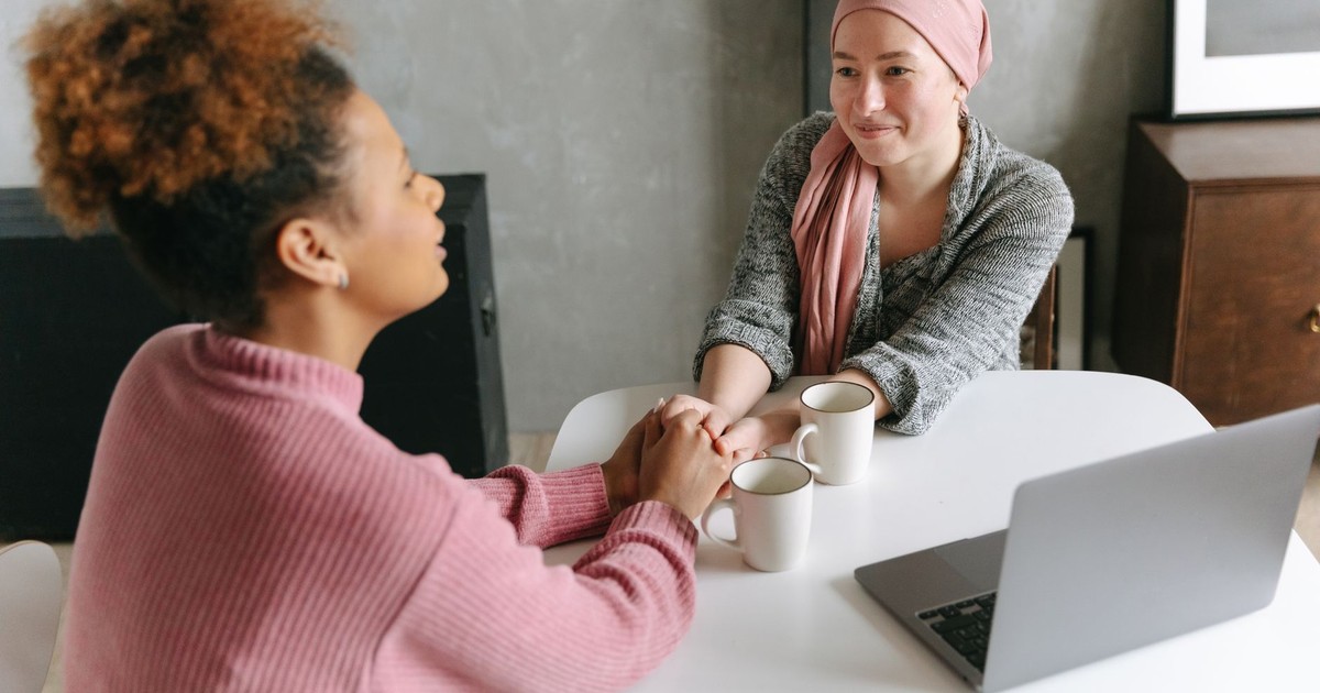 Two women sharing a supportive conversation over coffee indoors. One woman wears a head wrap.