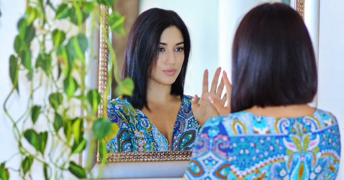 A woman in a vibrant patterned shirt gazes at her reflection in a mirror surrounded by green plants.