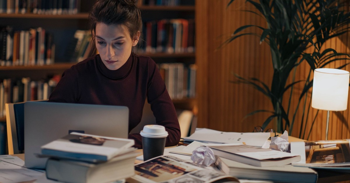 Woman working late at a desk filled with books, laptop, and coffee in a cozy library setting.