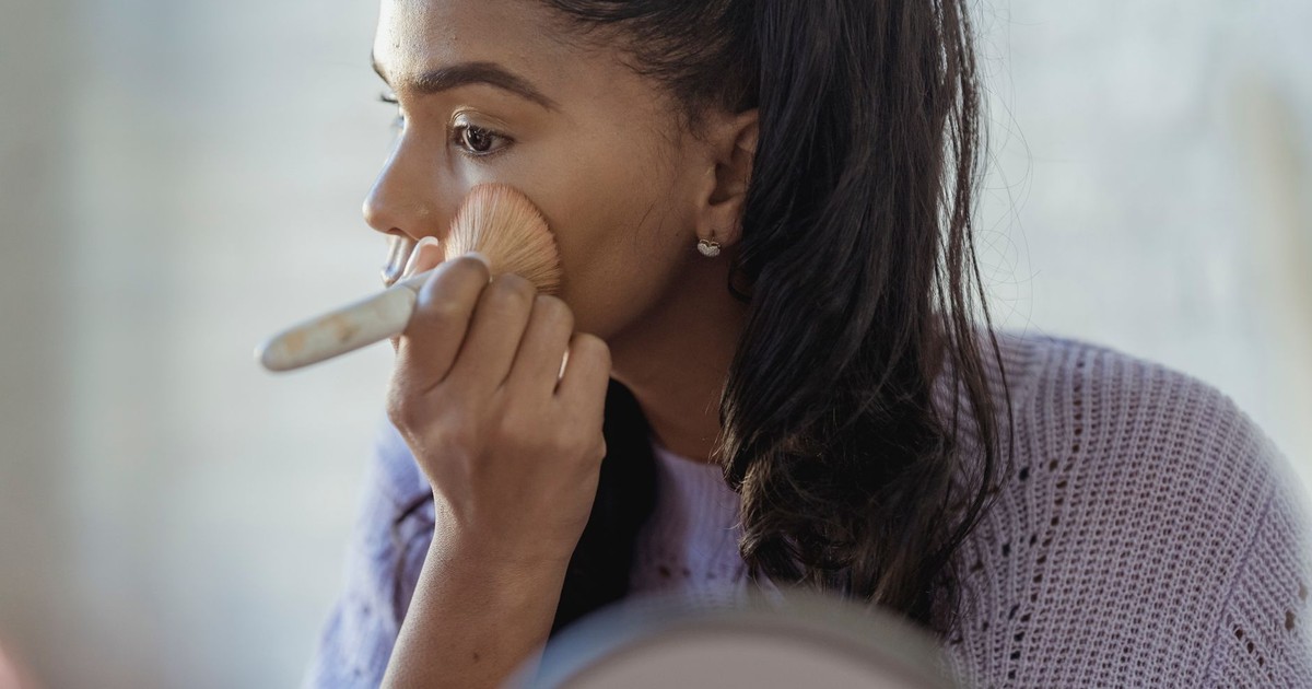 Concentrated crop African American young female spreading foundation cream on face with brush on blurred background
