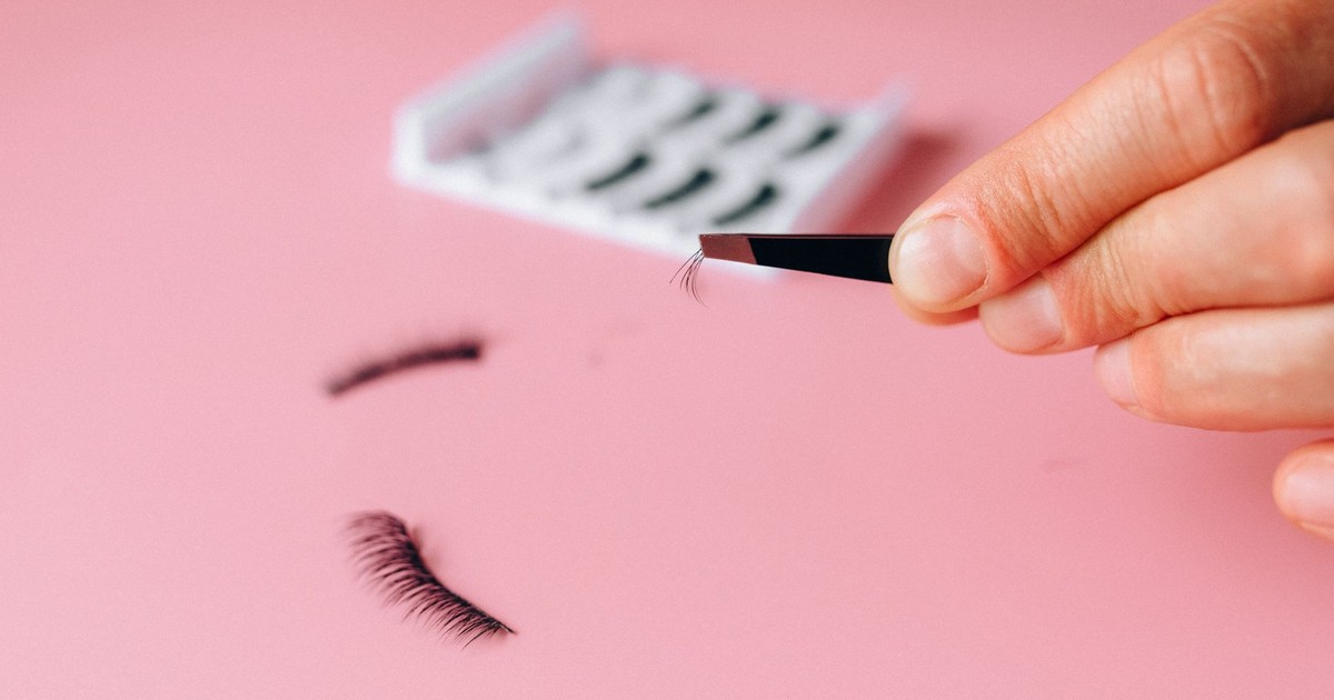 Close-up of applying false eyelashes with tweezers over a pink background.