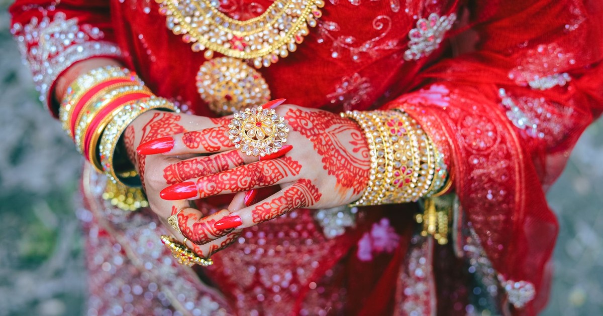 A close-up of a bride's hands adorned with red henna, gold jewelry, and a red dress, showcasing cultural elegance.