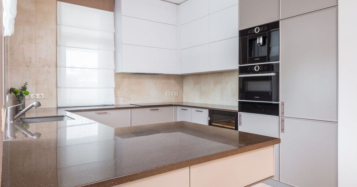 Contemporary kitchen with white and beige cupboards and built in appliances and cutting table laid with stone countertop and metal sink