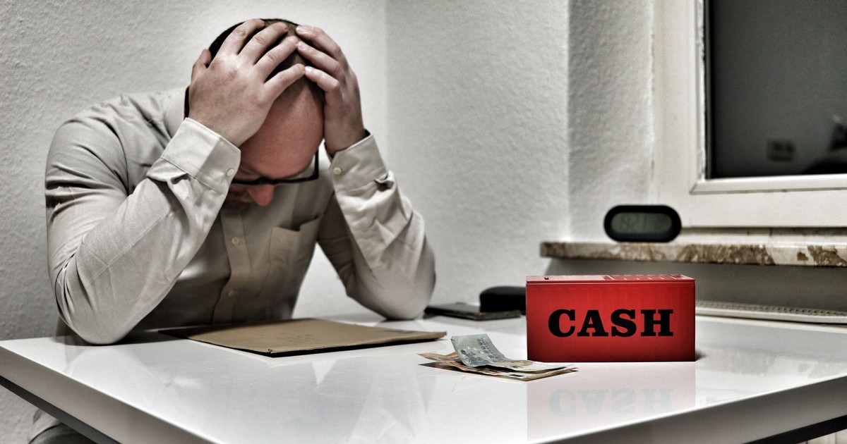 Bald man holding head in despair at desk with cashbox and money. Indoor office stress concept.
