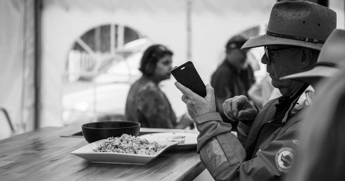 Black and white photo of an elderly man using a smartphone while dining indoors.