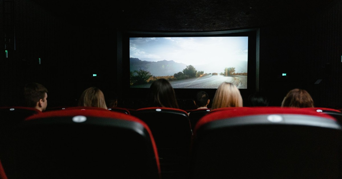 View from behind of people watching a movie in a cinema with red seats and a large screen.