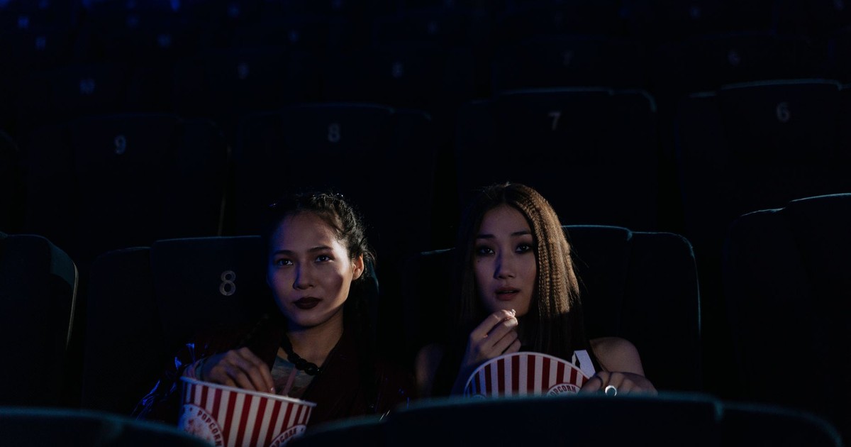 Two women sitting in a dark cinema, enjoying popcorn and watching a movie.
