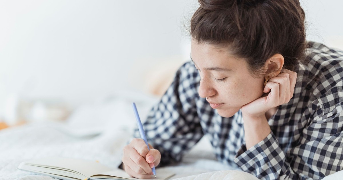 Young woman in checkered pajamas writing in a notebook while lying on a bed.