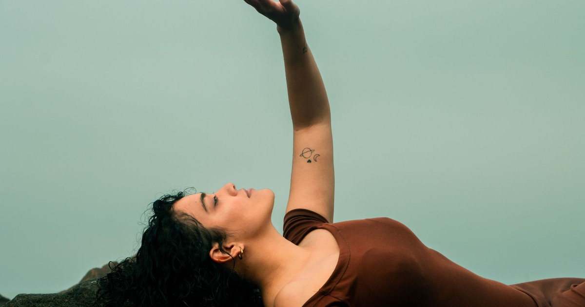 A woman in a brown dress relaxes outdoors in Lima, Peru, with her hand raised and a tattoo visible.