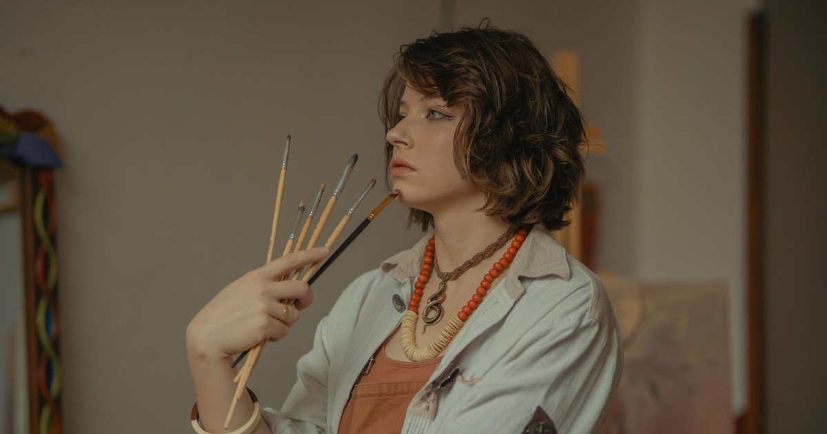 A young female artist stands in her studio, holding paintbrushes with a serene expression.