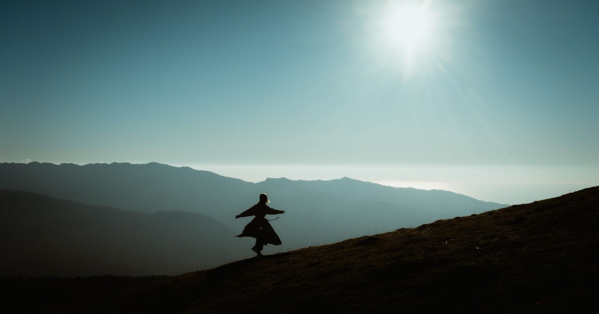 A serene silhouette of a person walking on a mountain slope under a bright sun and clear sky.